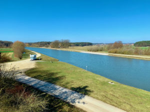 Blauer Himmel in der Natur, links ein Weg, rechts ein Fluss, ein Wohnmobil ist in der ferne zu sehen