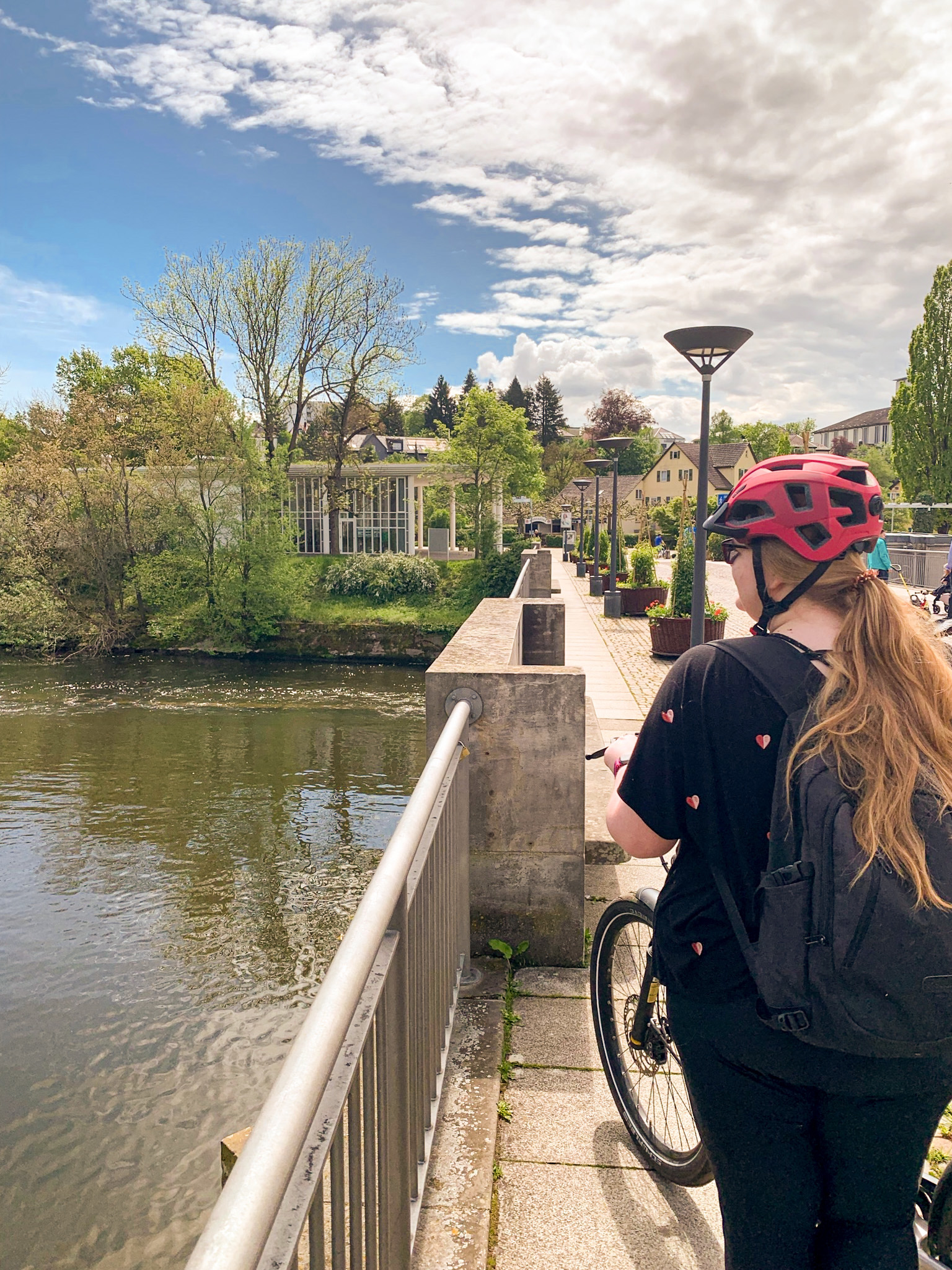 Fahrradtour Frau mit langen blonden Haaren von hinten und rotem Helm, auf einer Brücke stehend in die Ferne sehend, blauer Himmel mit wenigen Wolken, Bietigheim-Bissingen